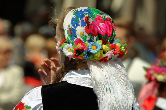 Młoda kobieta, blondynka, widziana z tyłu (g&oacute;rna część ciała), z włosami zakrytymi kwiecistą chustą z frędzlami, w białej koszuli z ludowym wzorem na rękaiwe, czarnym bezrękawniku, w tle rozmyty tłum