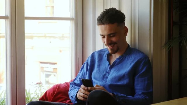Handsome bearded man with stylish hairstyle sitting near a window at home and using smartphone for chatting then thoughtful looking away smiling calm face online communication chat app type message