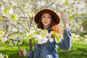 Young ginger hair woman in hippie gypsy jeans jacket, brown hat near white tender blossoming tree. Fairy dreamlike mood of spring or summer. Beautiful romantic lady smiling. Freedom travel lifestyle.