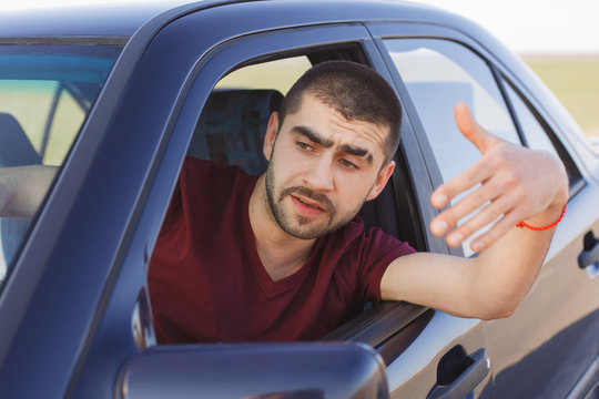 Portrait of handsome male driver waves from car window, being stuck in traffic jam, feels nervous and worried, looks into distance, notices accident on road. People, troubles on road, journey