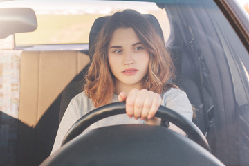 Photo of attractive female driver sits in car, teaches to drive, being inexperienced, has thoughtful expression. Woman in transport, stuck in traffic jam, thinks about something during long journey