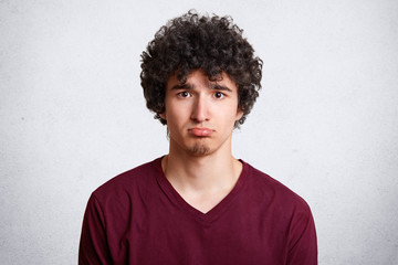 Fototapeta na wymiar Headshot of upset attractive male curves lower lip, being dissatisfied with negative news, being sad and disappointed, poses against white concrete wall. Gloomy teenager discontent with something