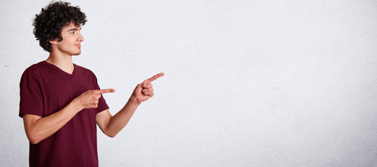 Horizontal portrait of handsome curly teenager has crisp dark hair, wears casual clothing, stands sideways and indicate with fore fingers at blank copy space for your promotional text or advertisement