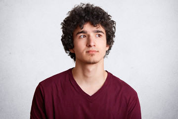 Obraz premium Close up shot of handsome curly teenager man looks pensively upwards, being deep in thoughts, ponders about future plans, isolated on white studio background. People, youth and lifestyle concept