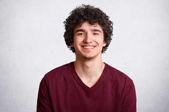 Portrait of happy male with broad smile, has curly hair, pleasant smile, dressed casually, stands against white background. Delighted attractive teenager rejoices having holidays after studying