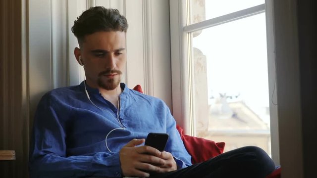 portrait of young worried caucasian man thoughtful using smartphone mobile with white headphones resting near window sunny day upset bad mood trouble internet connectivity issues home wifi device 3G