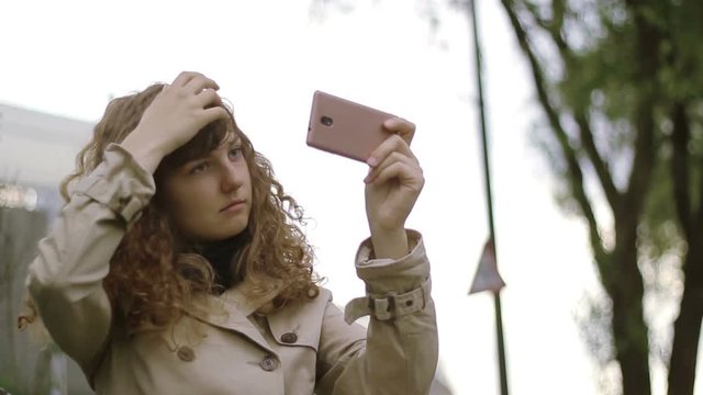 Pretty woman arranging her hair on the street with the help of a Smartphone.