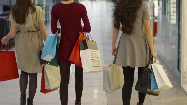 Rear view of three young women carrying paper bags with purchases when walking along shopping mall hallway
