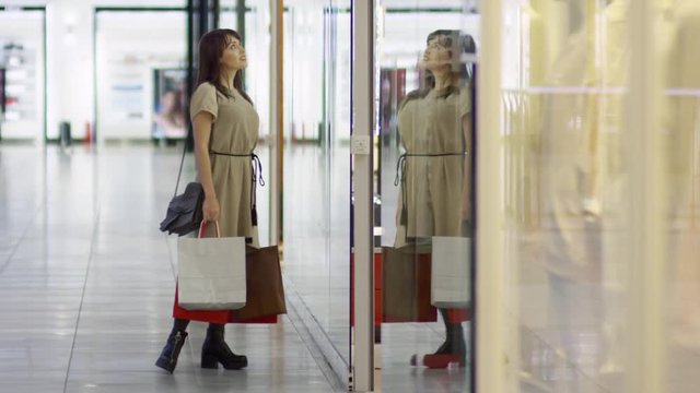 Tracking shot of happy female customer with shopping bags standing by display window and looking at outfits presented there