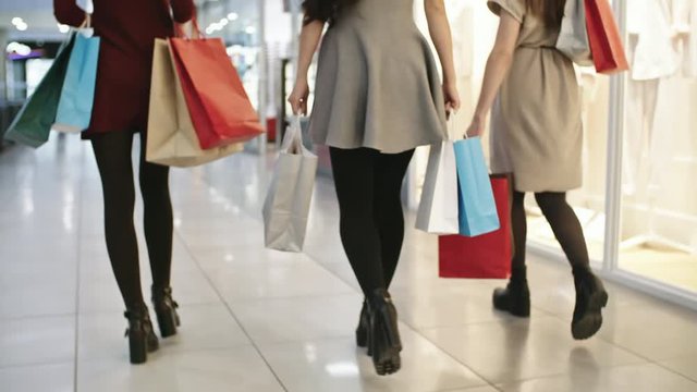 Tilt up shot of three young women carrying paper bags with purchases and looking into display windows when spending time together in shopping mall