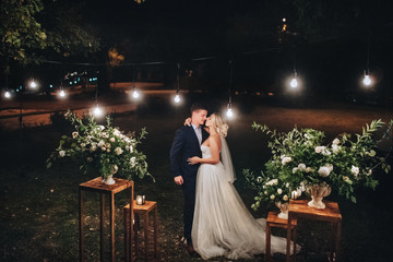 Stylish newlyweds stand at night against the backdrop of chic decorations and flowers, electric...
