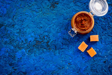 Caramel sauce in glass jar near caramel cubes on blue background top view copy space