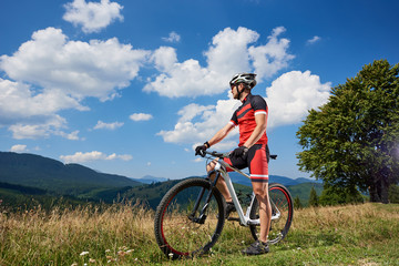 Fototapeta na wymiar Young athletic tourist biker standing with bike on grassy valley enjoying beautiful view of distant Carpathian mountains on bright sunny day. Active lifestyle and extreme sport concept.