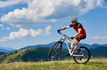Fototapeta na wymiar Young athletic sportsman tourist biker in professional sportswear turning on one wheel of bike on mountain hill on bright blue sky background. Active lifestyle and extreme sport concept.