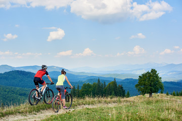 Back view of athlete couple cyclists in professional sportswear and helmets cycling cross country bikes on mountain trail on summer day in the Carpathians. Active lifestyle and outdoor sport concept
