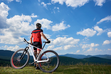Naklejka na ściany i meble Back view of young athletic professional tourist biker standing at bike on top of hill, enjoying beautiful view of distant mountains on bright summer day. Active lifestyle and extreme sport concept.