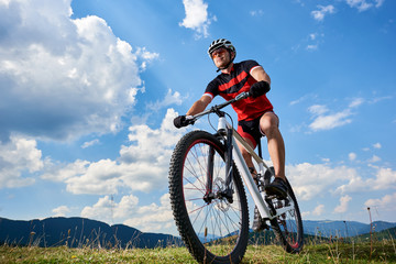 Fototapeta na wymiar Close-up front view of young athletic sportsman cyclist in professional sportswear riding a bike on distant mountains and bright blue summer sky background. Active lifestyle and extreme sport concept.
