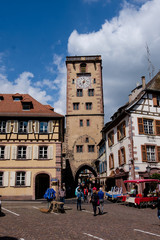 people walking along cobbled street bellow the clock tower and between beautiful half-timbered houses in Ribeauville, Alsace.