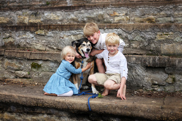 Three Cute Blonde Children Hugging Pet German Shepherd Dog Outside at Park