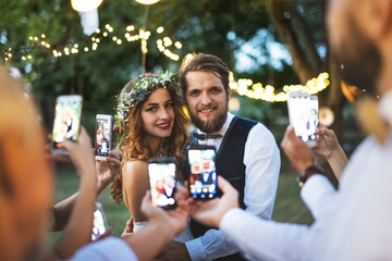 Guests with smartphones taking photo of bride and groom at wedding reception outside. - obrazy, fototapety, plakaty