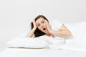 Fototapeta premium Calm young brunette woman lying in bed with white sheet, pillow, blanket on white background. Surprised beauty female spending time in room. Rest, relax, good mood concept. Copy space advertisement.