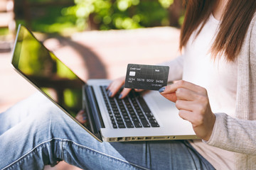 Naklejka premium Close up female hands on keyboard. Woman sitting on bench holding credit card, working on modern laptop pc computer in city park in street outdoors. Mobile Office. Online shopping, business concept.