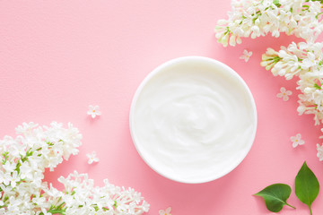 Opened white jar of natural herbal cream for women on pastel pink background. Beautiful branches of white lilac and petals. Care about clean and soft face, hands, legs and body skin. Fresh flowers.
