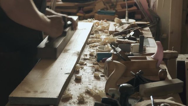 Skilled craftsman carpenter whittles workpiece of wooden board with jack-plane in a workshop. Making handmade furniture.