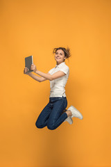 Image of young woman over orange background using laptop computer or tablet gadget while jumping.