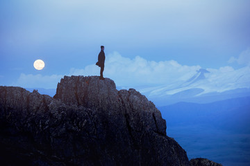Lonely man at evening stands on cliff against mountains and moon