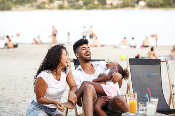 Young mixed race family sitting and relaxing at the beach on beautiful summer day.Parents making...