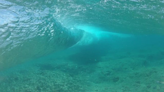SLOW MOTION, UNDERWATER: Empty barrel wave rolling over the beautiful reef in sunny Pacific. Unrecognizable surfer sits on his surfboard at line up while a big crystal clear tube wave rushes by him.