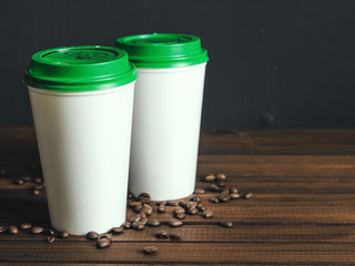 two white plastic coffee cups with a green lid on a wooden table with scattered coffee beans, copy space