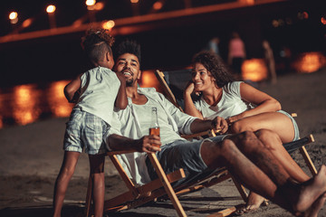 Fototapeta na wymiar Young mixed race family sitting and relaxing at the beach on beautiful summer night.They sitting on sun beds and drink beer.