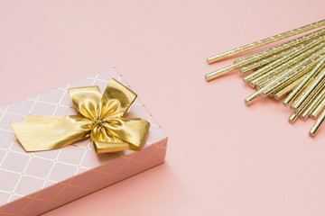 Female gift box with golden ribbon and cocktail accessories on punchy pastel pink. Copy space.