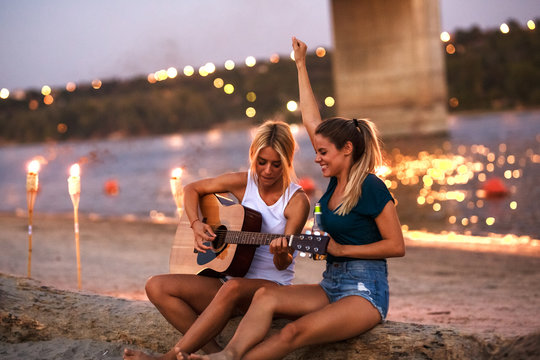 Two young female friends sitting by the river,singing and playing guitar.Joying in summer evening .