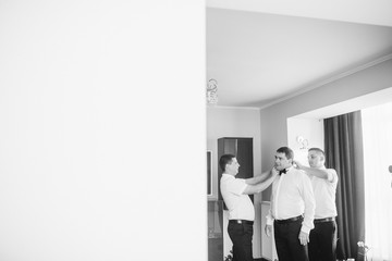Stylish handsome groom posing while groomsman putting on shirt and tying bow, helping him to get ready in the morning. wedding preparation. 