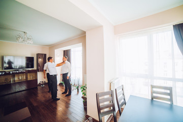 Stylish handsome groom posing while groomsman putting on shirt and tying bow, helping him to get...