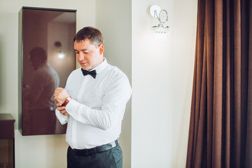 Fototapeta premium Stylish handsome groom posing while groomsman putting on shirt and tying bow, helping him to get ready in the morning. wedding preparation. 