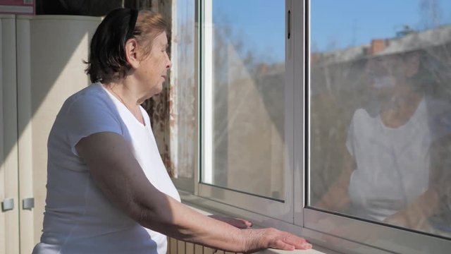 An Old Woman Stands By The Window And Looks Outside In Sunny Weather