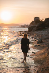 Back view shot of young fit woman with blonde hair in black beach dress walking along rocky beach near the beautiful sea. Sunset