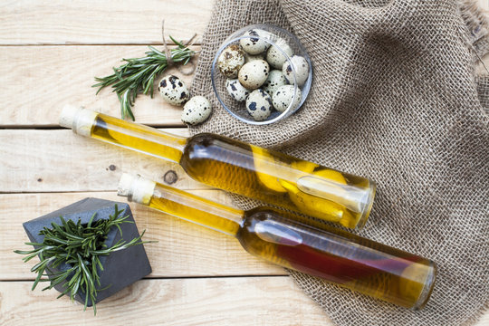 Two bottles of olive oil with lemon and pepper lying on a light wooden background. Next is a glass jug with quail eggs, a bunch of fresh rosemary lies.