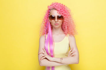 Fashion, style of youth. A beautiful girl in sunglasses with an incredible curly pink hairdo in a summer dress posing with her hands on her chest on a yellow background.