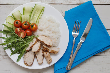 Portion of white rice, chicken breast, red cherry tomatoes, arugula leaves and cucumber slices served on white plate with blue napkin, fork and knife. Healthy and balanced eating
