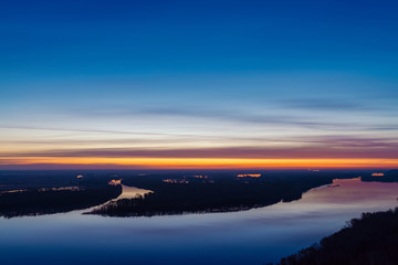 Beautiful river with big island with trees under predawn sky. Bright yellow stripe in picturesque cloudy sky. Early blue sky reflected in water. Colorful morning atmospheric image of majestic nature.