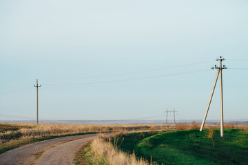 Fototapeta na wymiar Atmospheric landscape with power lines in green field with dirt road under blue sky. Background image of electric pillars with copy space. Wires of high voltage above ground. Electricity industry.