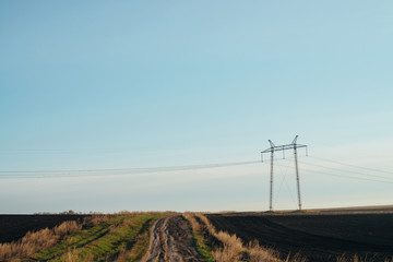 Obraz premium Atmospheric landscape with power lines in green field with dirt road under blue sky. Background image of electric pillars with copy space. Wires of high voltage above ground. Electricity industry.