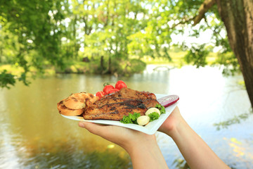 Kobieta trzyma w dłoniach grilowane mięso, pieczywo i warzywa na tle jeziora.