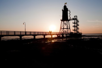 Silhouette eines Paares, das im Sonnenuntergang auf dem Seesteg vor dem Leuchtturm Obereversand in Dorum-Neufeld, Deutschland, spazieren geht