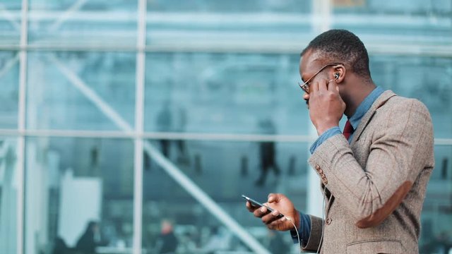 African American businessman listens to the music and dances while he walks near the office building with a smartphone. Concept: office worker, rest, advertising